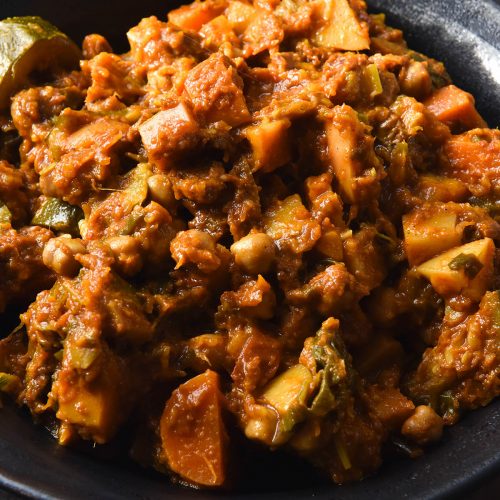 A close up of a dark ceramic bowl filled with FODMAP friendly vegetable tagine. The bowl sits atop a dark grey backdrop