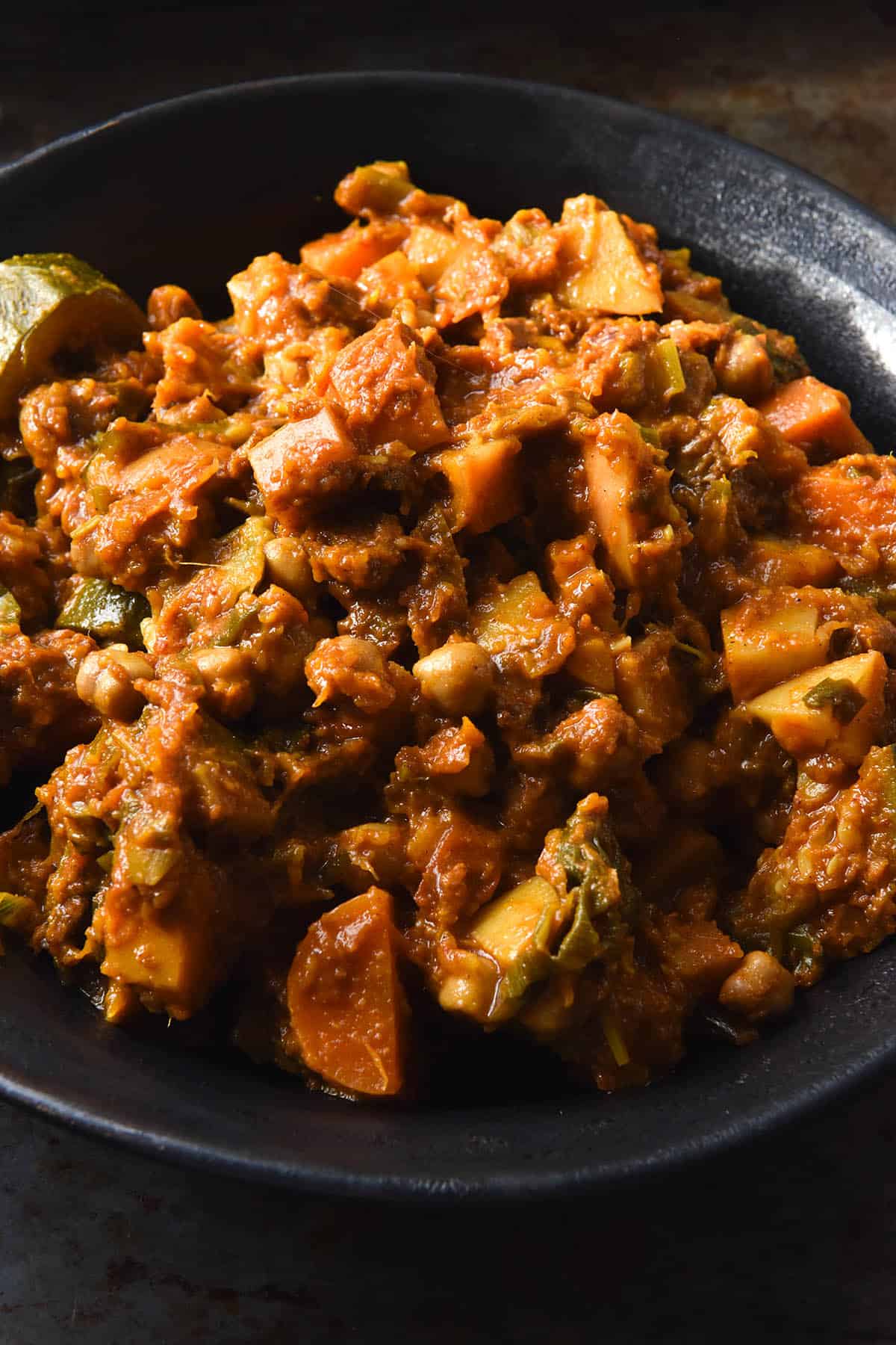 A close up of a dark ceramic bowl filled with FODMAP friendly vegetable tagine. The bowl sits atop a dark grey backdrop