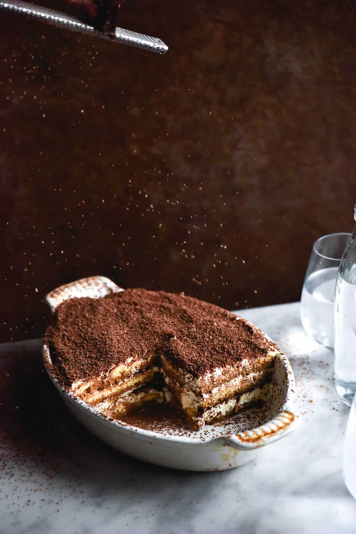 An aerial, side on image of a gluten free Tiramisu in a white baking dish. The tiramisu sits atop a white marble table against a rust coloured backdrop. Shards of chocolate sprinkle down from the top of the image onto the Tiramisu. Glasses of water sit to the right of the image, creating filtered light across the table.