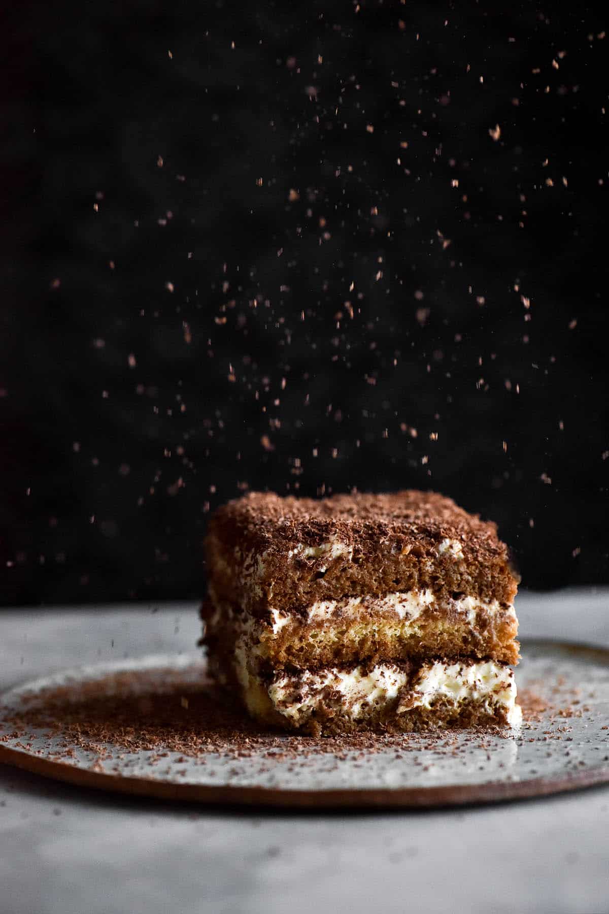 A single slice of gluten free, lactose free Tiramisu sits atop a small white ceramic plate atop a white marble table. The tiramisu slice sits against a black background, which contrasts against the gratings of chocolate falling down from the top of the image.