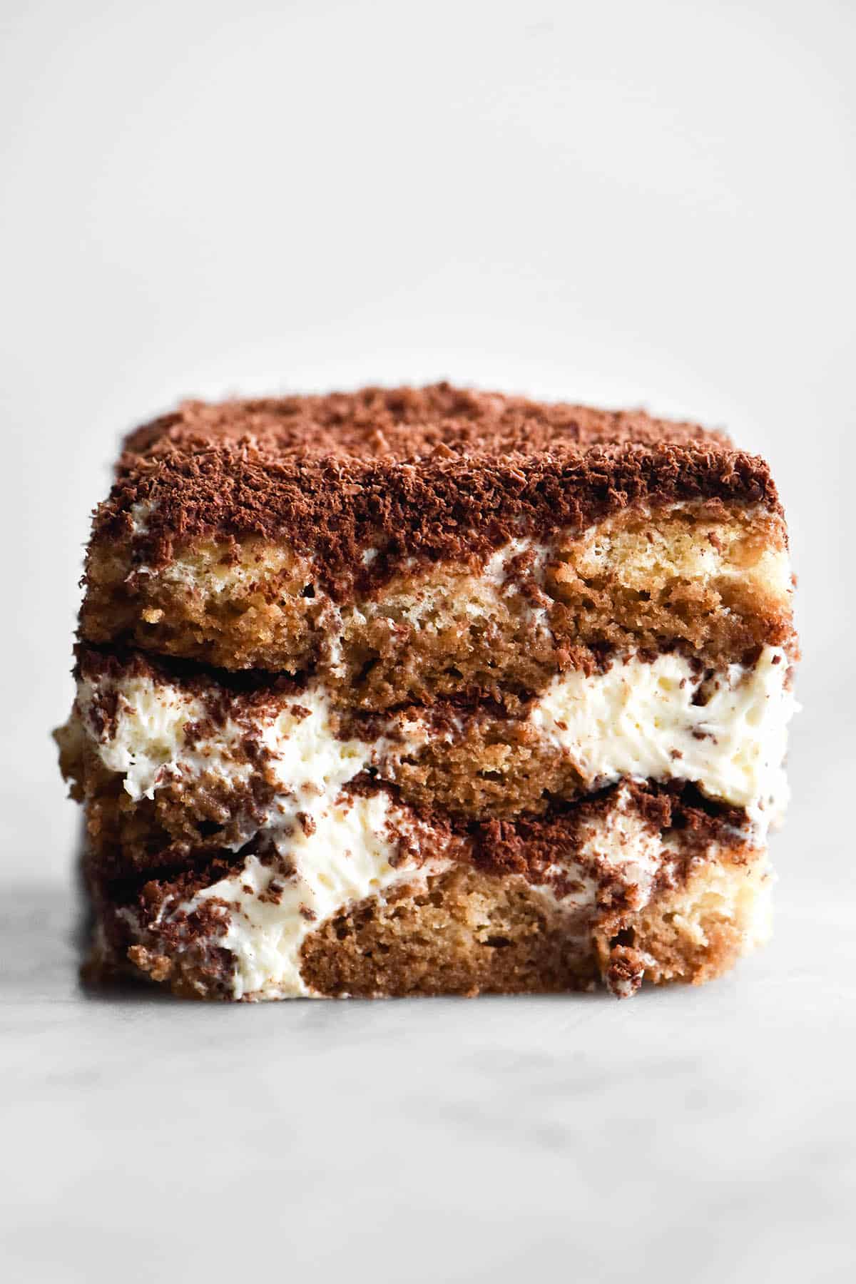 An side on macro image of a tall slice of gluten free Tiramisu sitting atop a white marble table against a white backdrop.