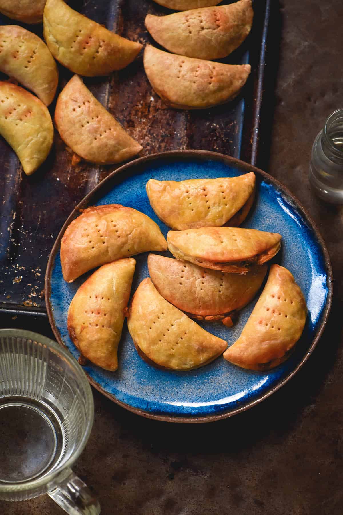An aerial image of a blue plate of gluten free empanadas on a rusty auburn backdrop. More empanadas sit on a baking tray above the plate, and a water jug sits to the bottom left of the plate.
