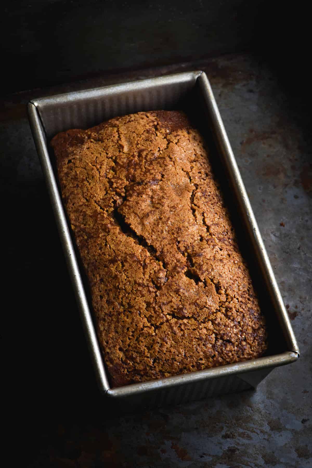 A moody, aerial image of a loaf of gluten free pumpkin bread. The pumpkin bread is golden brown and topped with finishing sugar for a crackly top. It sits in a metal loaf pan atop a dark grey metal background. 