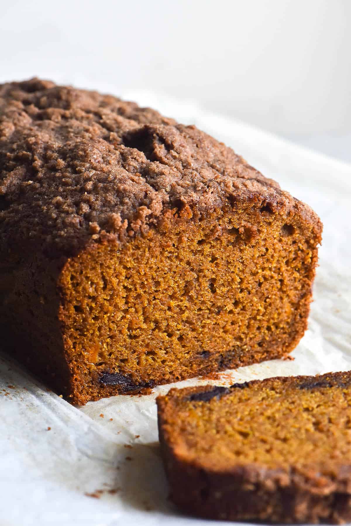 A side on, airy light photo of a loaf of gluten free pumpkin bread with a crumble topping. The loaf has a slice removed, revealing the bright orange crumb and a few studs of dark chocolate.