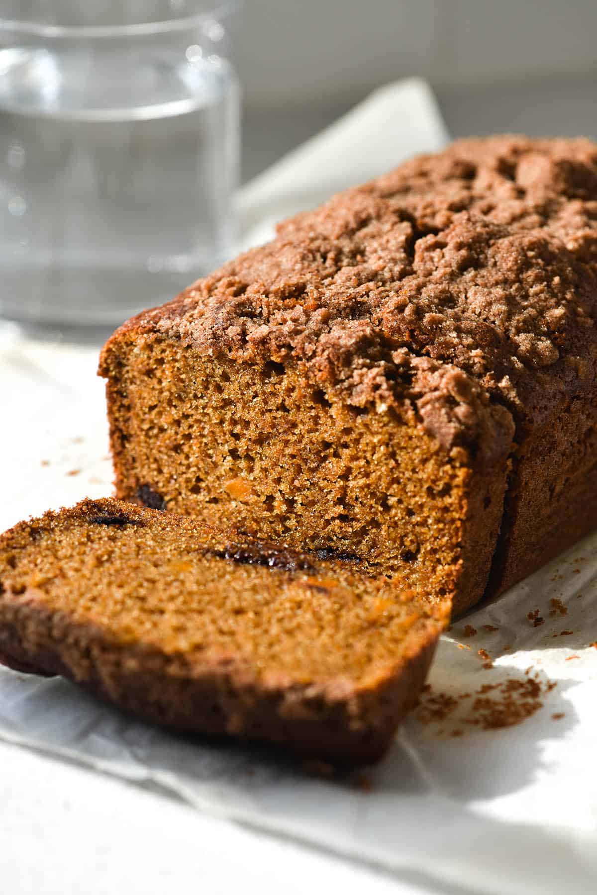 A side on macro image of a gluten free pumpkin bread atop a white countertop. The first slice lays flat so that the bright orange crumb is revealed, as are the chocolate chips. A glass of water sits in the background of the image. 