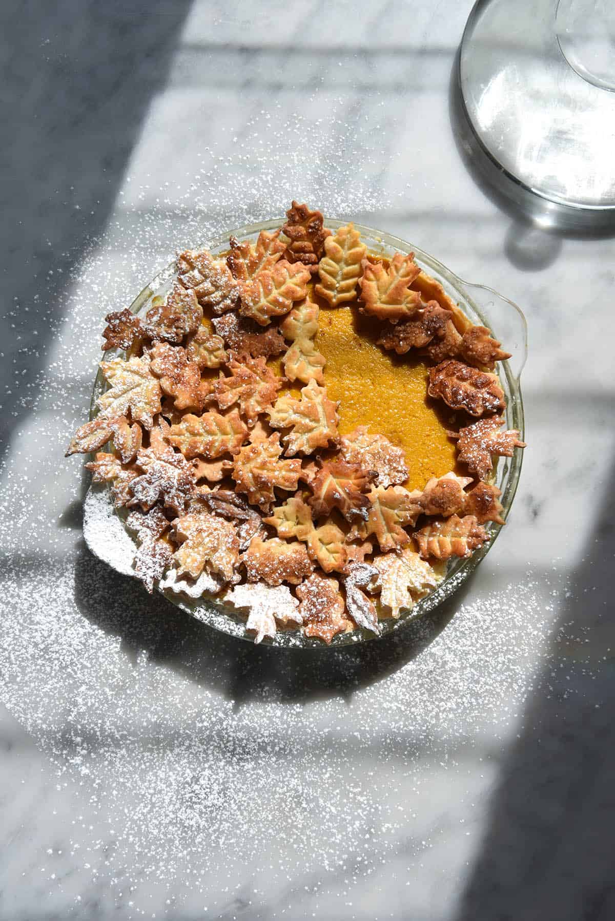 An aerial image of a gluten free pumpkin pie topped with ornamental pastry leaves and a sprinkling of icing sugar. The pie sits atop a white marble table that sits in heavily contrasted sunlight. A jug of water sits to the top right of the image, further amplifying the sun and shadow. 