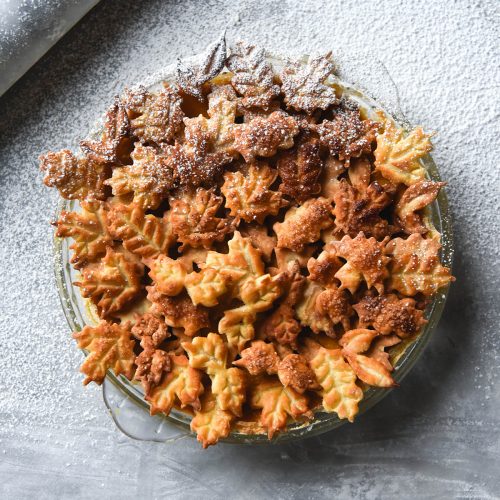 An aerial view of a gluten free pumpkin pie topped with ornamental pastry leaves on a white marble table. The table is sprinkled with icing sugar and a white marble rolling pin sits in the top left of the image.