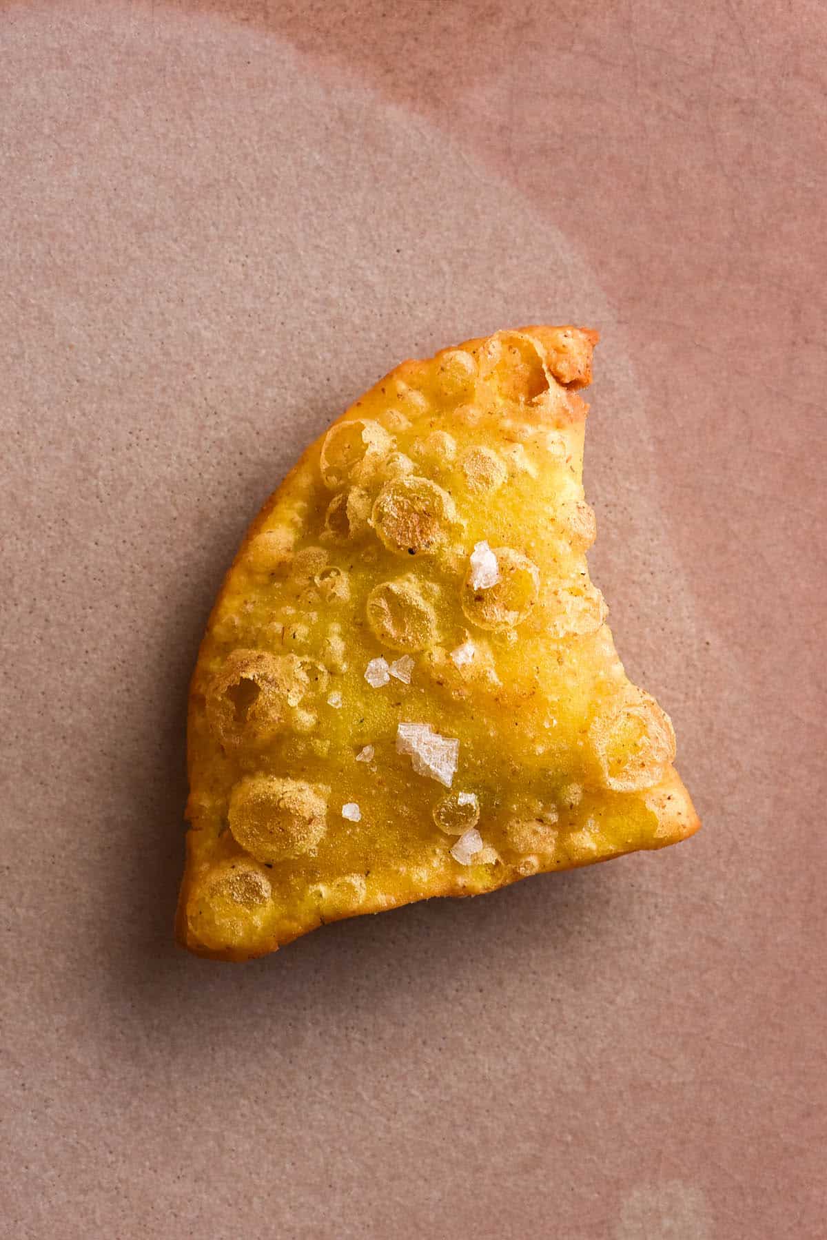 An aerial close up of a golden brown gluten free samosa topped with sea salt flakes. The samosa sits on a textured pale pink ceramic plate.