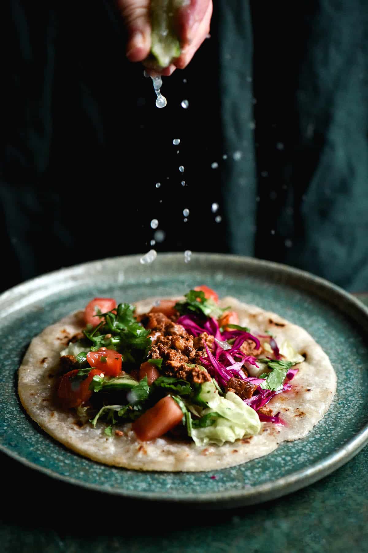 A side on photo of a gluten free tortilla topped with vegetarian taco mince and salsa. The tortilla sits atop an green plate on an olive green table, set against an olive green linen backdrop. A hand extends down from the centre top of the image to squeeze lime onto the tortilla. The droplets of lime juice are frozen in frame on the way down.