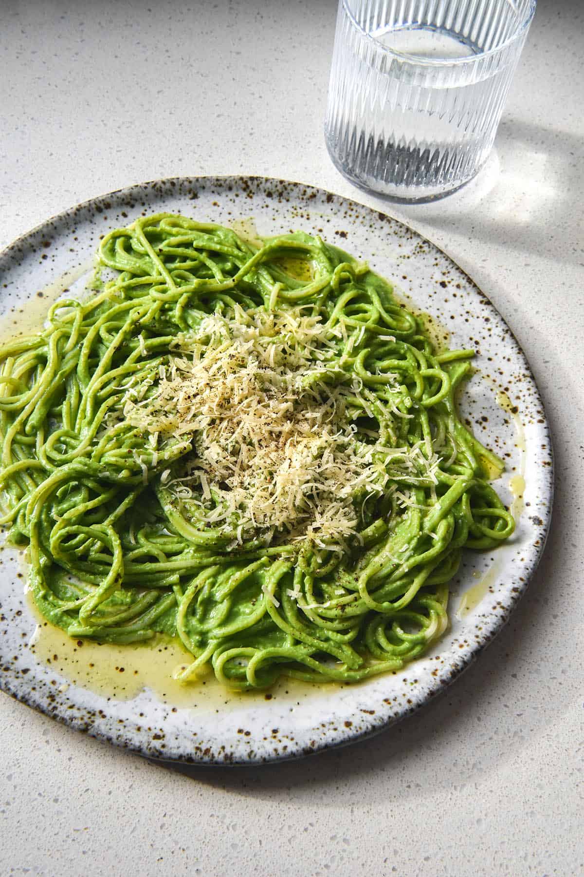A side on sunlit view of a white ceramic plate topped with spaghetti in a green protein pasta sauce. The spaghetti is topped with grated parmesan, freshly cracked pepper and garlic infused oil. It sits atop a white stone benchtop and a glass of water sits to the right of the plate.