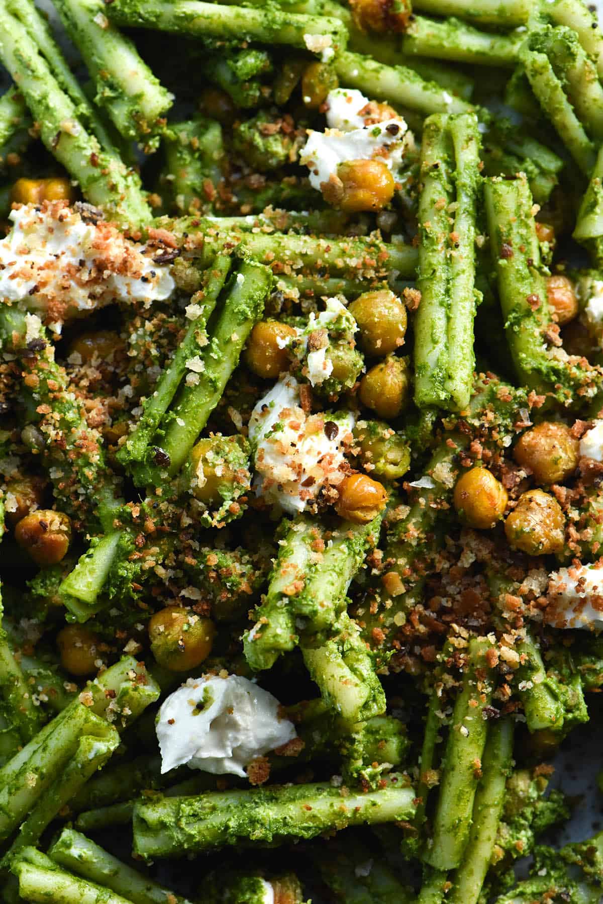 A close up aerial view of a plate of gluten free casarecce smothered in green protein pasta sauce, topped with some crispy chickpeas, breadcrumbs and burrata. 