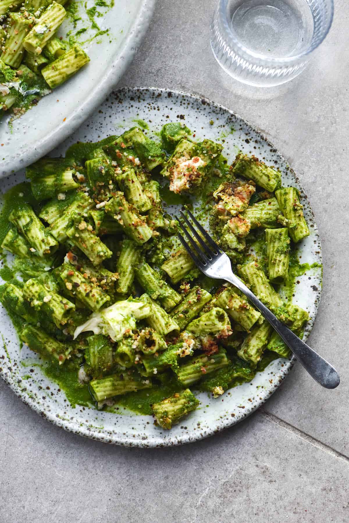 An aerial photo of a plate of rigatoni smothered in green protein pasta sauce and topped with bread crumbs and burrata. The central plate is a white speckled ceramic plate with a fork to the right of the plate. A second plate sits in the top left corner, both atop a light grey stone backdrop.