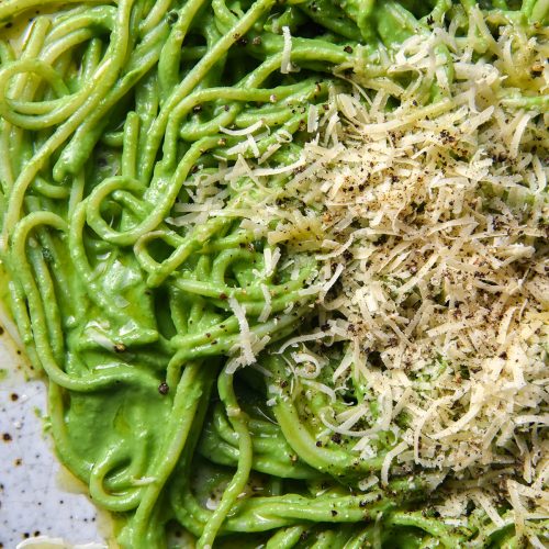 A close up aerial view of a white speckled ceramic plate topped with spaghetti in green protein pasta sauce. The spaghetti is then topped with freshly grated parmesan, black pepper and garlic infused oil.