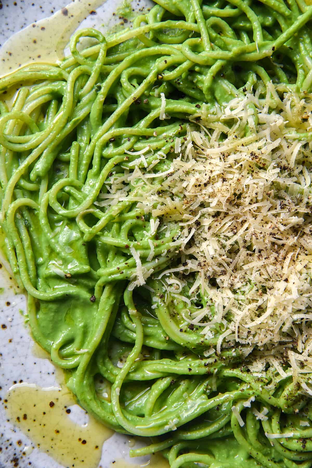 An aerial, close up view of a plate of gluten free spaghetti topped with green protein pasta sauce, grated parmesan, pepper and garlic infused oil. The spaghetti sits atop a white speckled ceramic plate. 