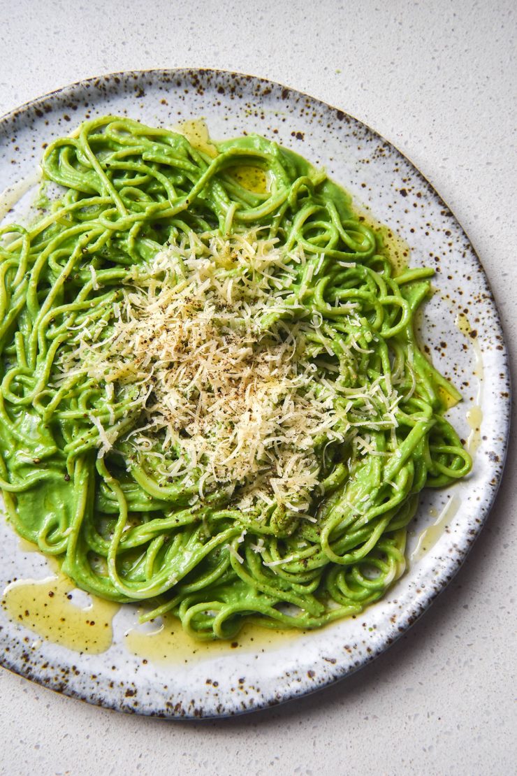 An aerial view of a plate of green protein pasta sauce smothered gluten free spaghetti. The spaghetti is then topped with grated parmesan, cracked pepper and garlic infused oil. It sits atop a white speckled ceramic plate on a white stone benchtop.