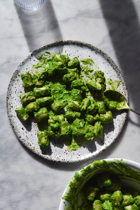 An aerial sunlit image of Low FODMAP kale pesto on gluten free potato gnocchi. The plate of gnocchi sits atop a white marble table and a glass of water creates a contrasting shadow across the image