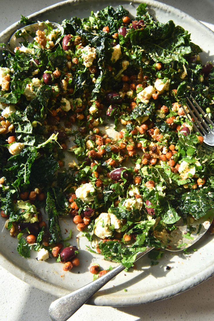 A sunlit close up photo of a kale, crispy chickpea, feta and raisin salad with a bold lemon, honey and mustard dressing. The salad sits atop a white ceramic serving plate against a white backdrop.