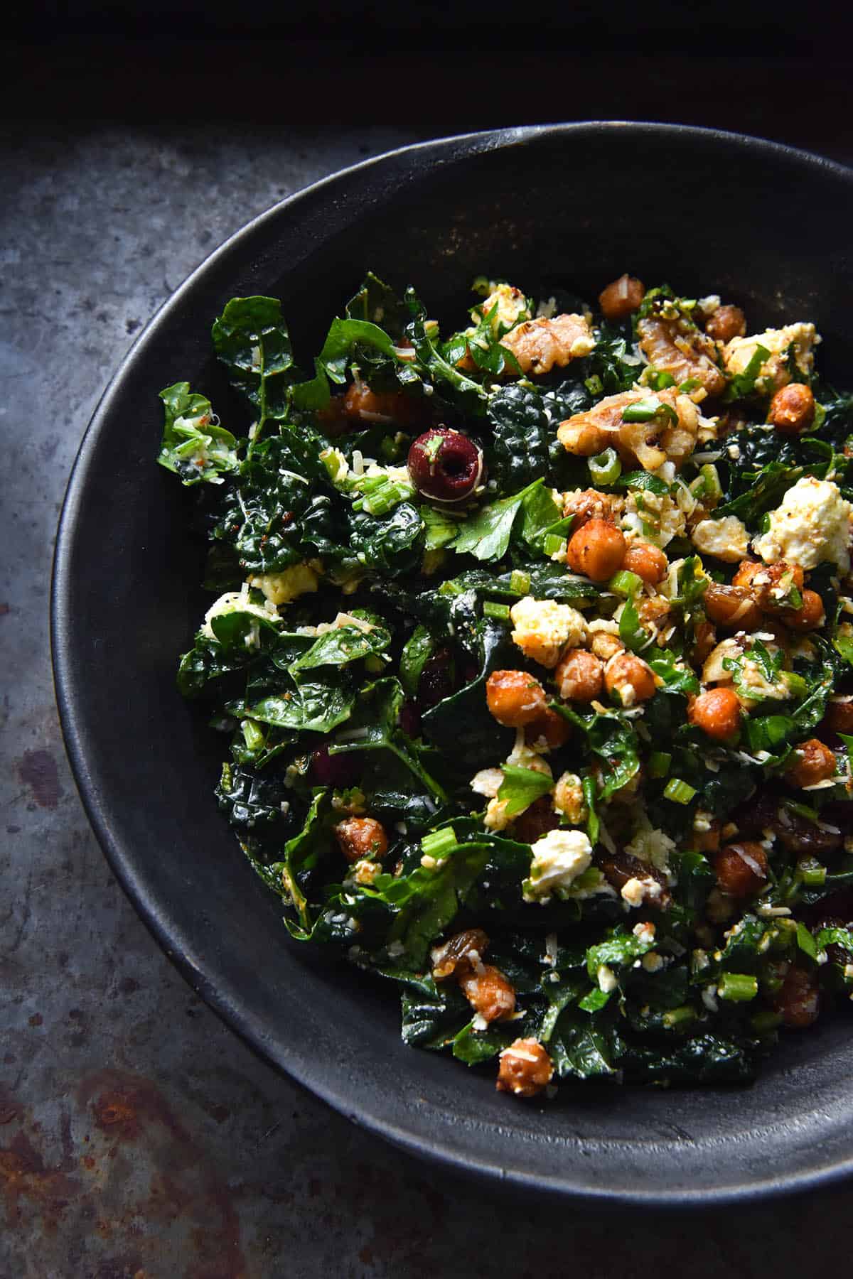 A darker image of a grey ceramic bowl filled with a kale, crispy chickpea, feta and raisin salad. The salad is piled into the bowl which sits atop a dark mottled grey background.