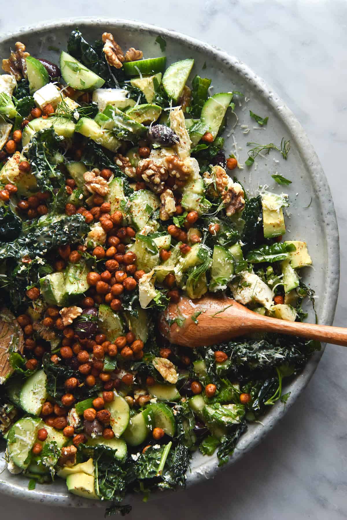 A close up aerial image of a kale salad with crispy chickpeas, cucumber, olives, walnuts, avocado and a zingy mustard herb dressing. The salad is topped with grated parmesan and sits atop a large white ceramic serving plate on a white marble table.