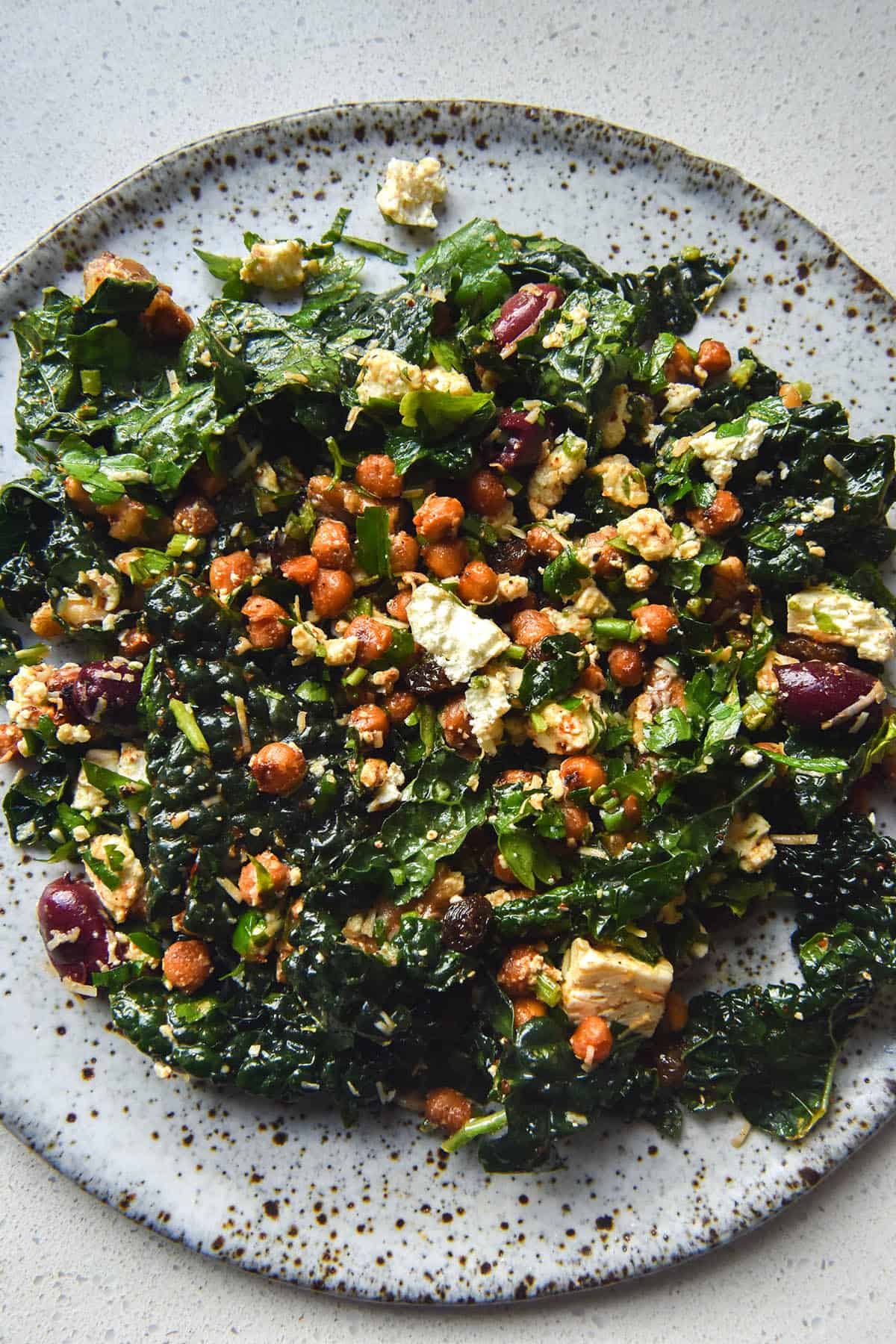 A close up aerial image of a white speckled ceramic plate topped with a kale, crispy chickpea, feta and raisin salad. The white speckled ceramic plate is in the centre of the image and sits atop a white benchtop.