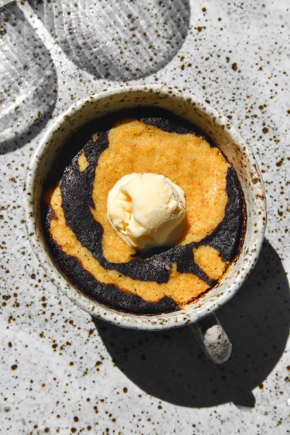 An aerial view of a gluten free pumpkin and chocolate swirl mug cake topped with a scoop of vanilla ice cream. The mug is a white speckled ceramic one that sits atop a white speckled ceramic plate. Two water glasses sit to the top left of the image and cast a pattern across the photo.