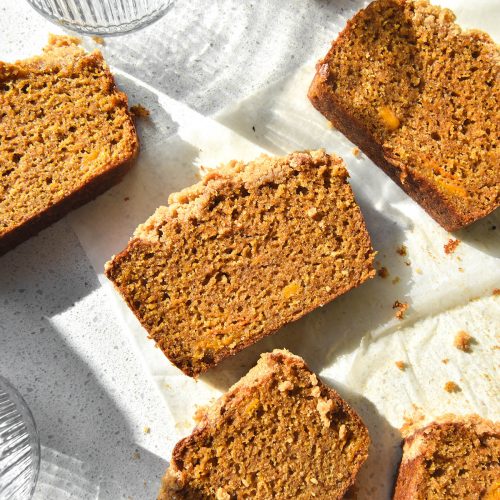 An aerial image of slices of gluten free pumpkin bread atop a white sunlit benchtop. The slices are casually arranged half on a piece of baking paper and half on the counter. Two textured water glasses sit to the left of the image and cast light and shadow across the slices of pumpkin bread.