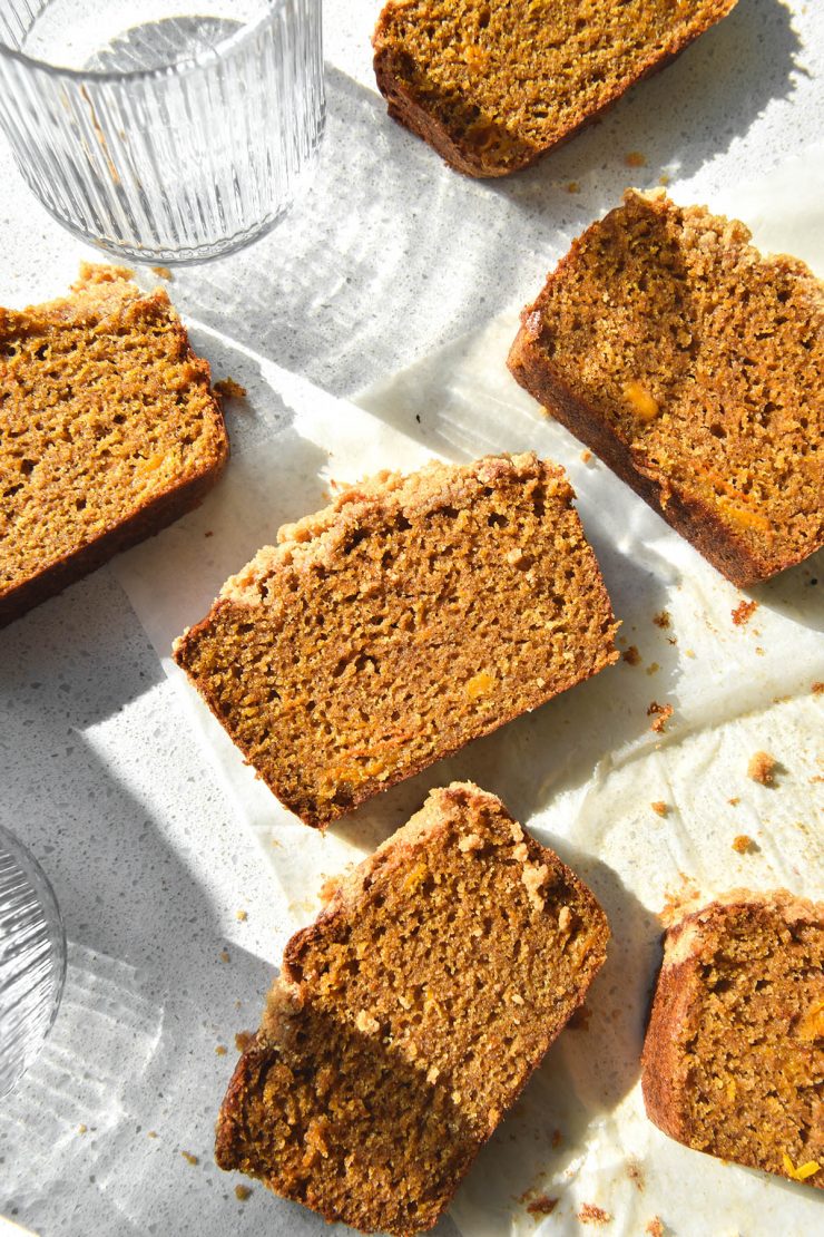 An aerial image of slices of gluten free pumpkin bread atop a white sunlit benchtop. The slices are casually arranged half on a piece of baking paper and half on the counter. Two textured water glasses sit to the left of the image and cast light and shadow across the slices of pumpkin bread.