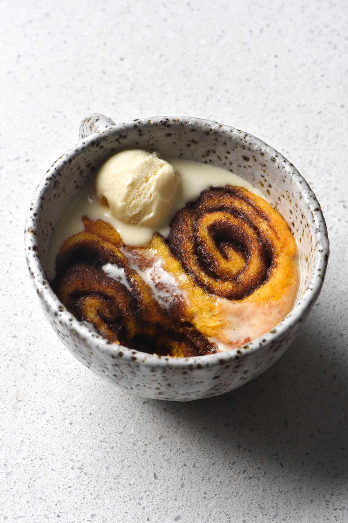 A close up aerial image of a gluten free pumpkin cinnamon scroll mug cake atop a white speckled counter. The mug is a white speckled ceramic mug and is filled with two pumpkin cinnamon scrolls, topped with melting vanilla ice cream. 