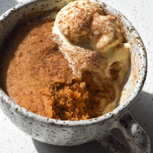 A side on view of a brightly lit gluten free pumpkin mug in a white speckled ceramic mug. The mug cake is topped with vanilla ice cream with a sprinkling of cinnamon sugar. A spoonful of the mug cake has been eaten from the bottom right corner, revealing the orange crumb beneath.