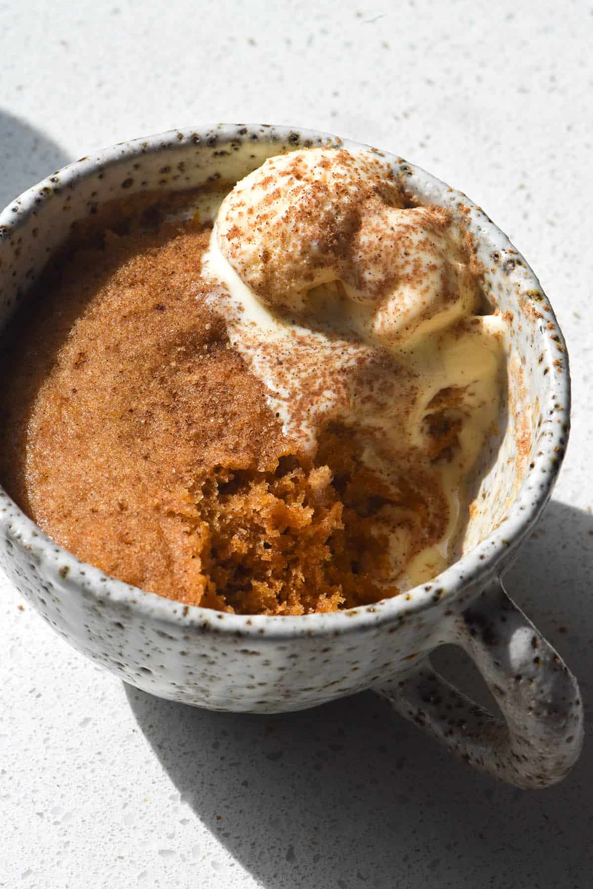 A side on close up view of a gluten free and vegan pumpkin mug cake. The cake sits in a ceramic white speckled mug against a white speckled countertop. It is topped with melting ice cream and a dusting of cinnamon sugar. A big spoonful has been taken out of the front of the mug cake, revealing the orange crumb below the surface