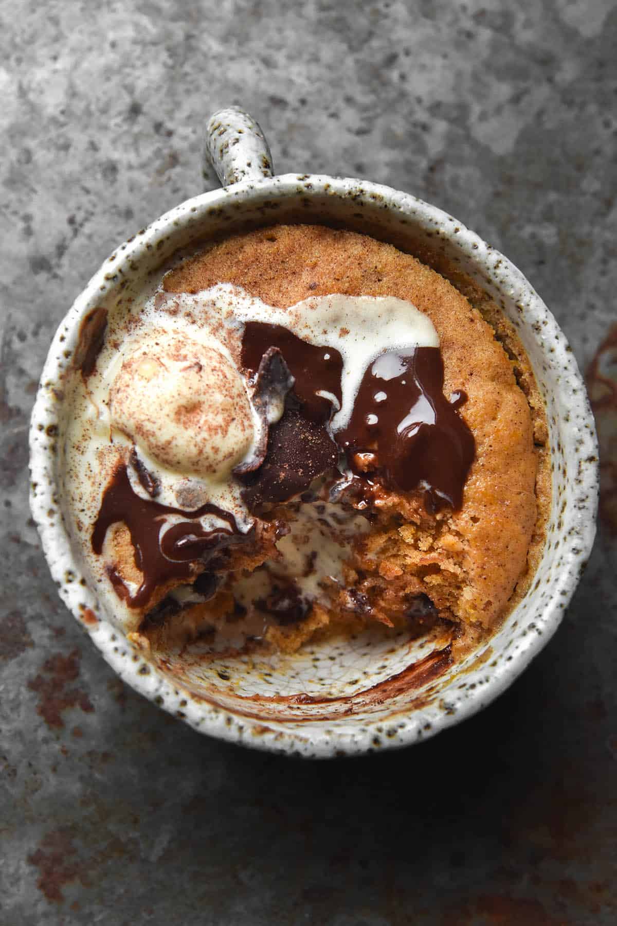 An aerial view of a gluten free pumpkin mug cake in a white speckled mug on a mottled grey backdrop. Melting vanilla ice cream and chocolate sauce mingle on the top of the cake and melt into the crevice left behind from a spoonful of cake being removed. 