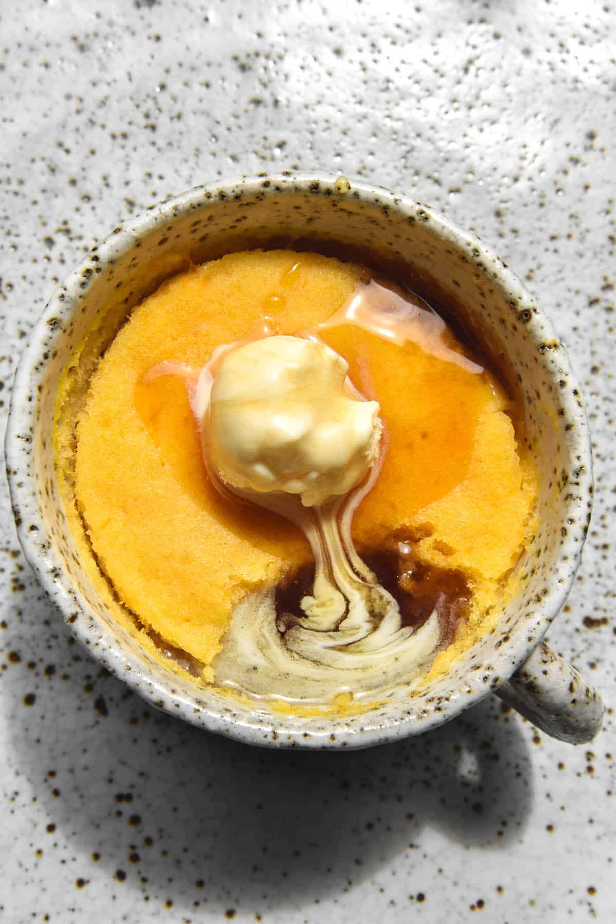 A gluten free pumpkin mug cake in a white speckled ceramic mug on a white speckled ceramic plate. The mug cake is a bright orange, bathed in sunlight. It is topped with melting ice cream and maple syrup that pools together in the hole where a spoon of mug cake has been eaten. 