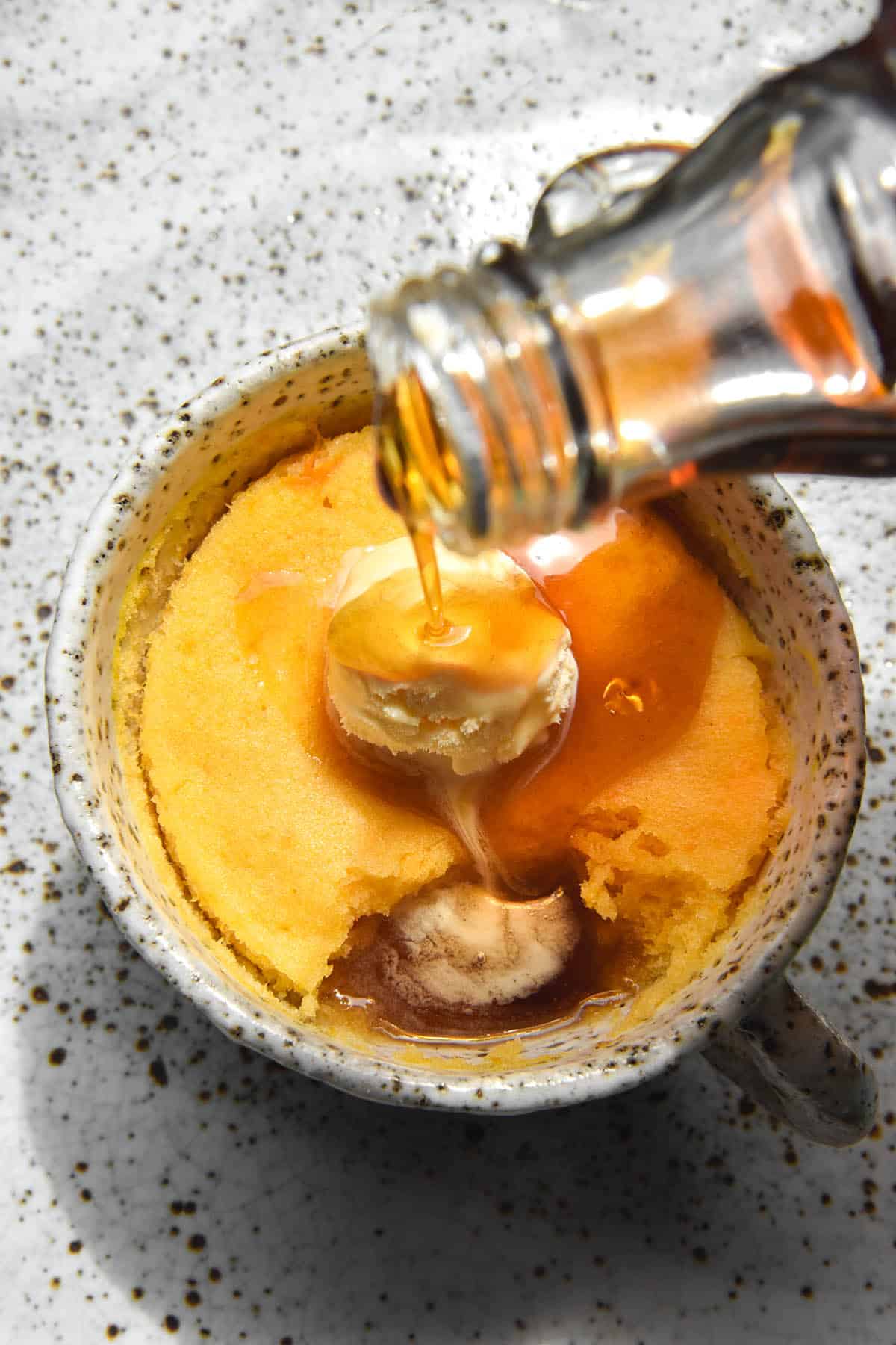 A sunlit image of a gluten free pumpkin mug cake in a white speckled ceramic mug on a white speckled ceramic plate. The mug cake is topped with a scoop of vanilla ice cream and a bottle of maple syrup extends from the centre right of the image to pour down onto the ice cream. The maple syrup and melted ice cream meld together in a pool created by a spoonful of the pudding being removed. 