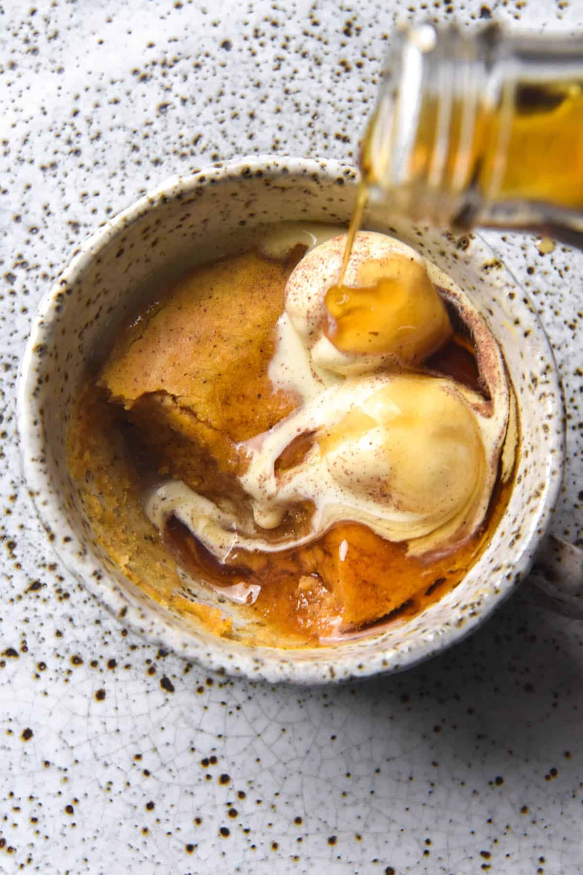 An aerial image of a gluten free pumpkin mug cake topped with vanilla ice cream, cinnamon sugar and being drizzled with maple syrup. The mug is a white ceramic speckled one, as is the plate it sits atop. The melting ice cream and maple syrup mingle in a pool where cake has been eaten out of the mug. 