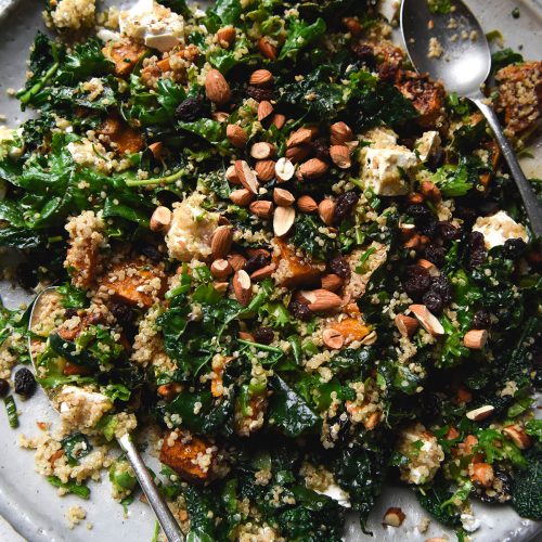 An aerial view of a white ceramic plate topped with a roasted pumpkin quinoa salad with feta, raisins, kale and almonds. The salad sits casually arranged on the serving platter, which sits atop a white benchtop. Two serving spoons sit on the dish on the left and right sides.