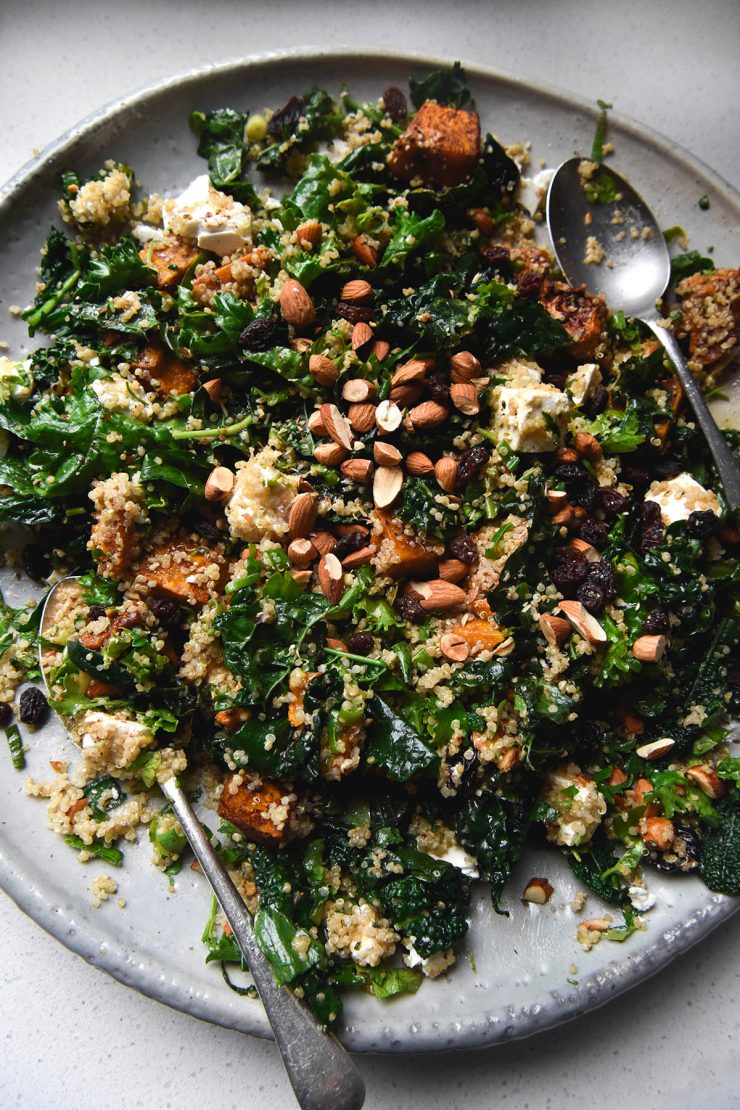 An aerial view of a white ceramic plate topped with a roasted pumpkin quinoa salad with feta, raisins, kale and almonds. The salad sits casually arranged on the serving platter, which sits atop a white benchtop. Two serving spoons sit on the dish on the left and right sides.