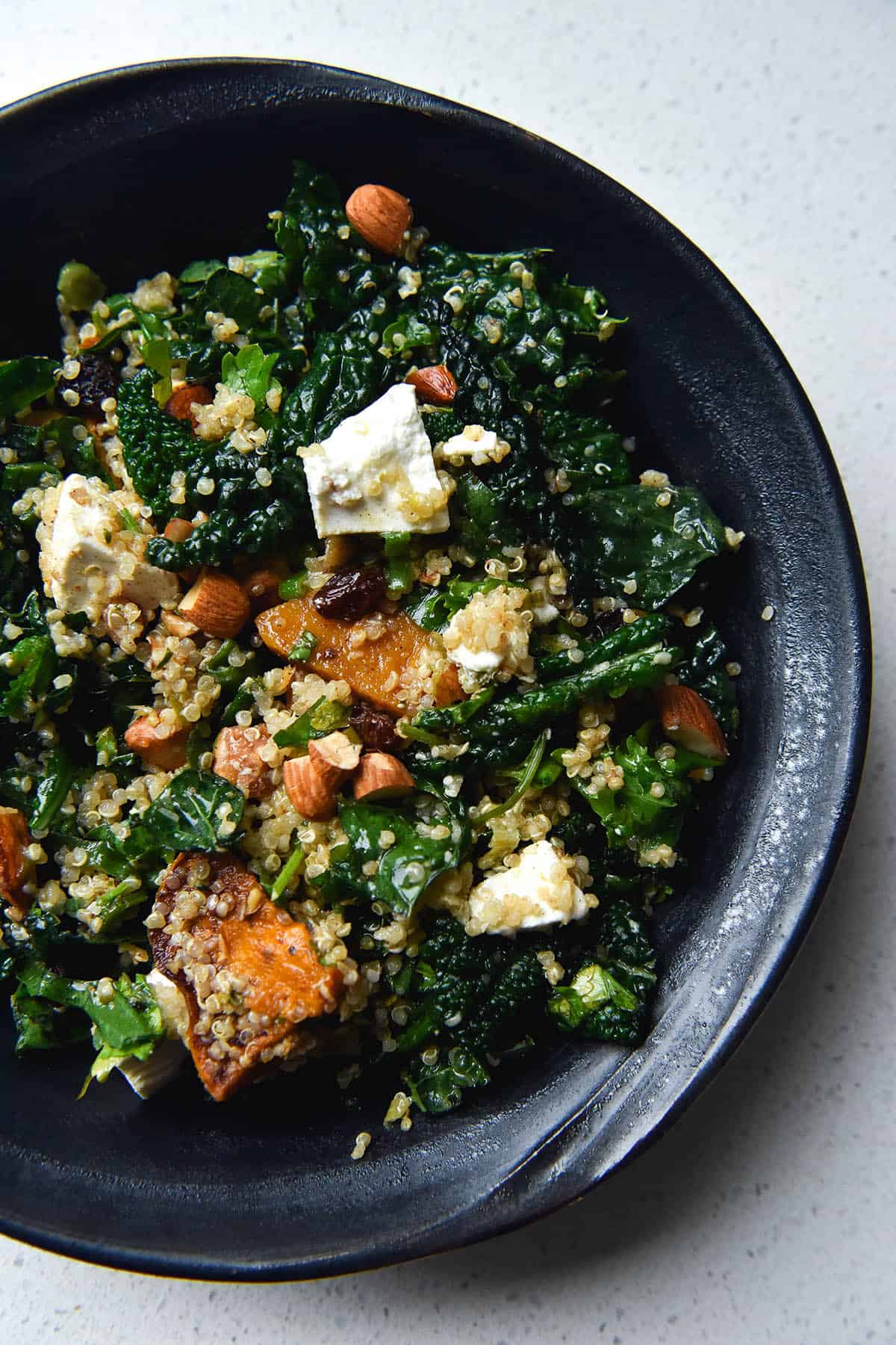 An aerial, close up view of a dark grey ceramic bowl filled with a roasted pumpkin, quinoa, kale, feta, almond and raisin salad. The bowl sits atop a white stone benchtop.