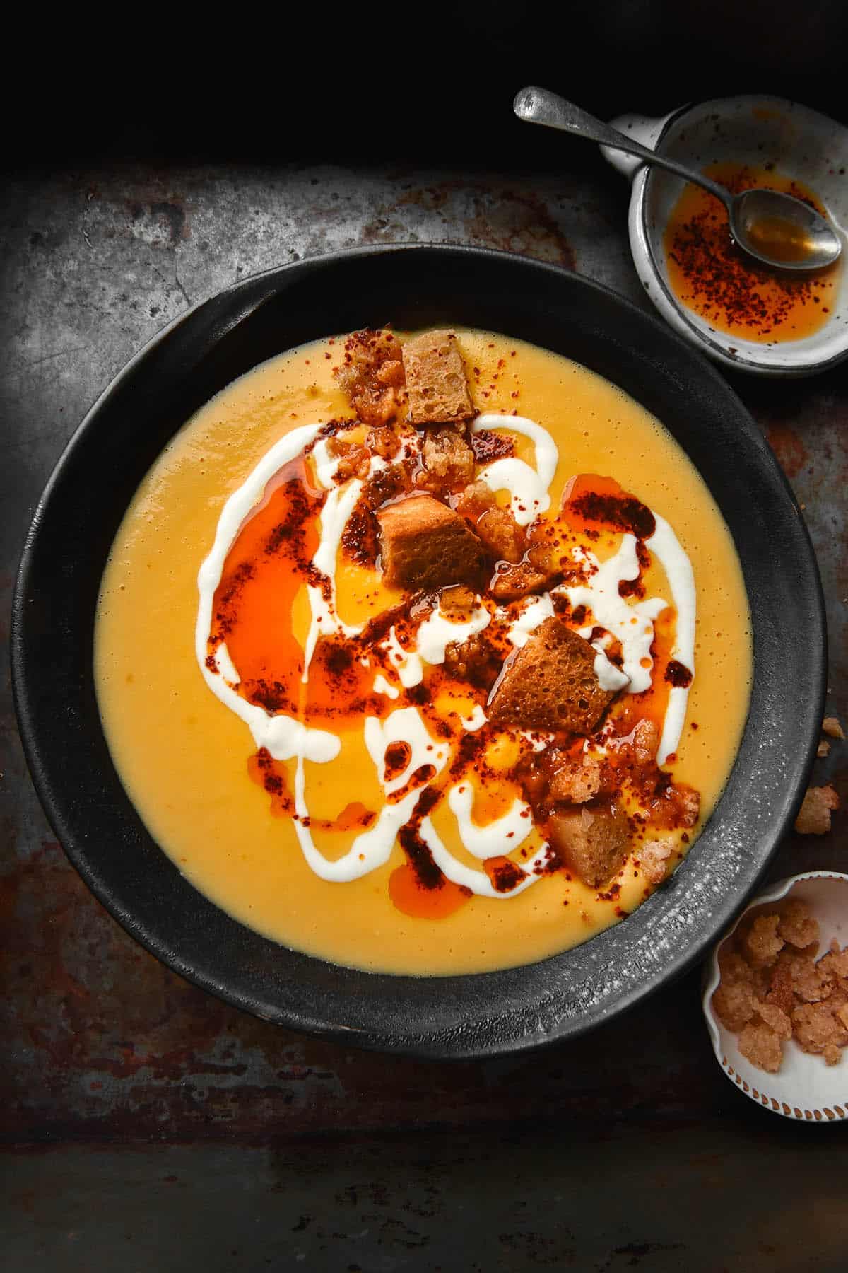 An aerial, moody photo of a dark grey ceramic bowl filled with pumpkin soup. The pumpkin soup is topped with thinned yoghurt, garlic infused chilli oil, garlic infused croutons and an extra sprinkle of Aleppo chilli. The bowl sits atop a dark grey backdrop. A small white ceramic dish filled with chilli oil and a spoon sits to the top right of the image. A small white ceramic bowl filled with extra garlic breadcrumbs sits to the bottom right of the image. 