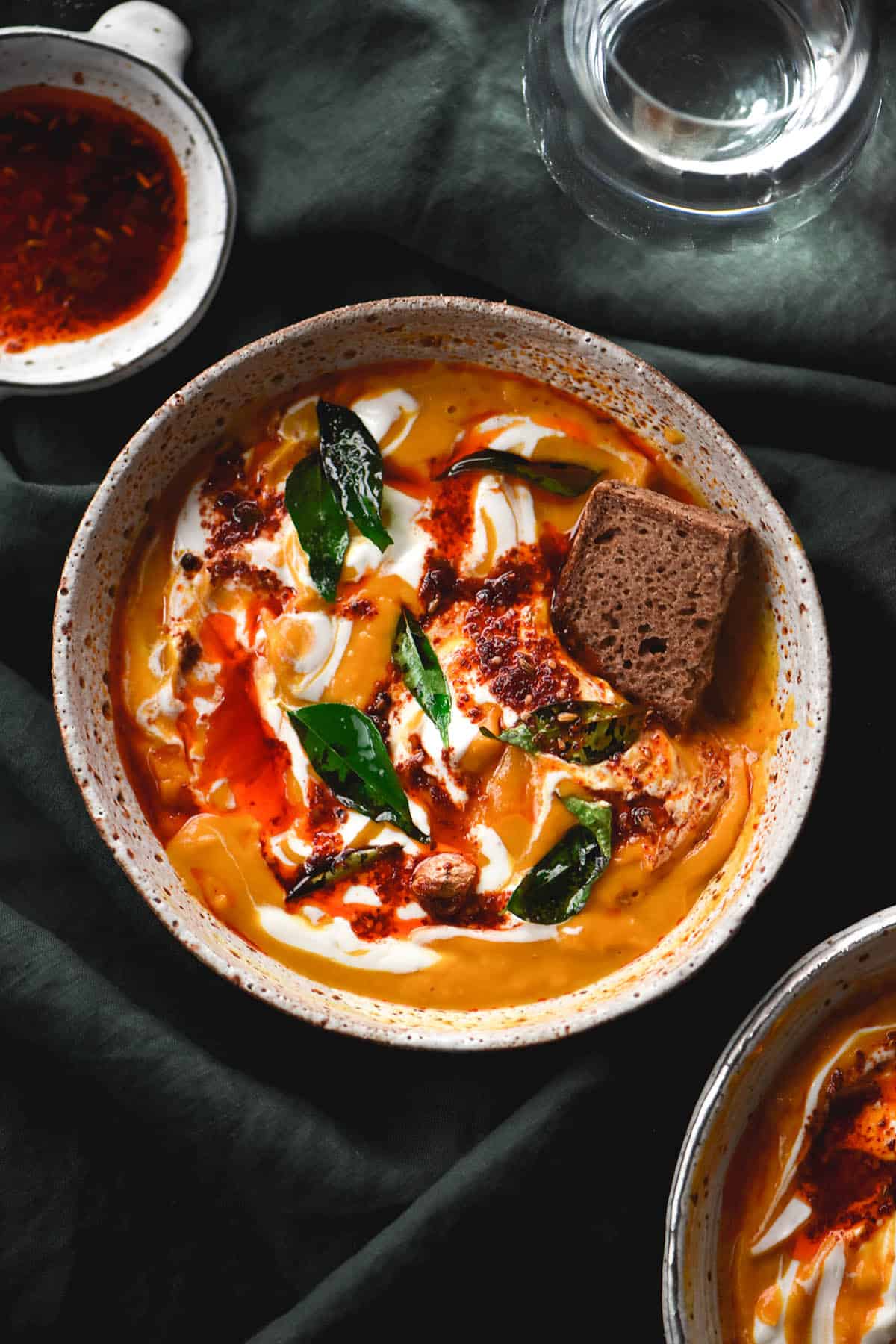 An aerial view of a white ceramic bowl filled with pumpkin soup sitting atop an olive green linen backdrop. The pumpkin soup is topped with cream, chilli oil, fried curry leaves and dukkah. A piece of dark brown sourdough dips into the bowl on the right side of the image. A small white ceramic dish of chilli oil sits to the top left, and a glass of water to the top right. A second bowl of pumpkin soup sits in the bottom right corner, just visible. 