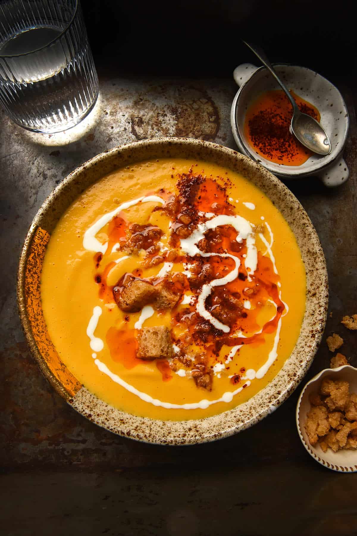 An aerial, moody photo of a speckled beige bowl filled with pumpkin soup. The pumpkin soup is topped with thinned yoghurt, garlic infused chilli oil, garlic infused croutons and an extra sprinkle of Aleppo chilli. The bowl sits atop a dark grey backdrop. A small white ceramic dish filled with chilli oil and a spoon sits to the top right of the image. A small white ceramic bowl filled with extra garlic breadcrumbs sits to the bottom right of the image. A glass of water sits to the top right of the image. 