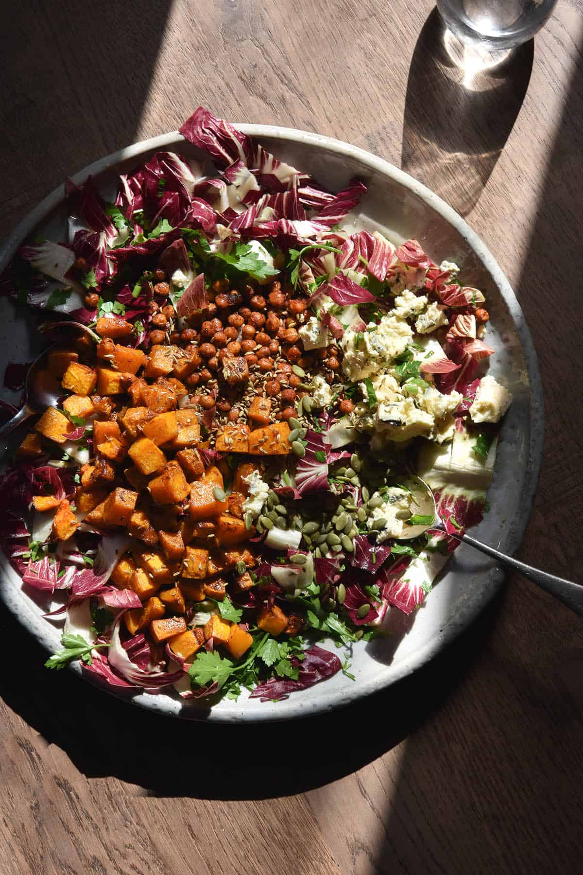 An aerial, sunlit view of a radicchio, blue cheese, smoky chickpea, roasted pumpkin and parsley salad on a white ceramic serving dish. The salad sits atop a medium brown wood backdrop in a central sliver of sunlight. The edges of the dish and salad are cast in shadow. 