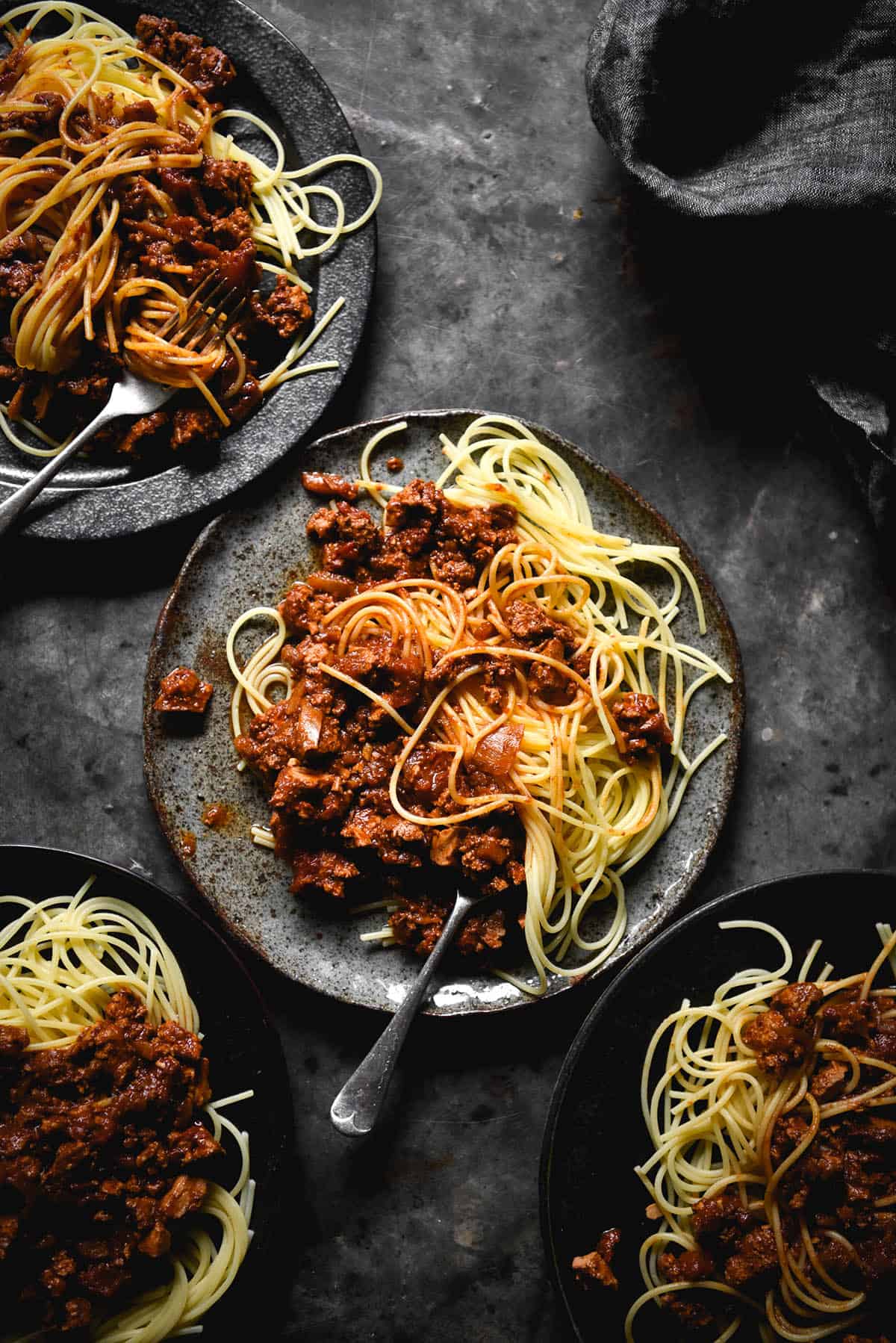 An aerial view of four plates of vegan bolognese atop gluten free spaghetti. Each plate is a different style dark colour ceramic and they sit atop a dark steel backdrop