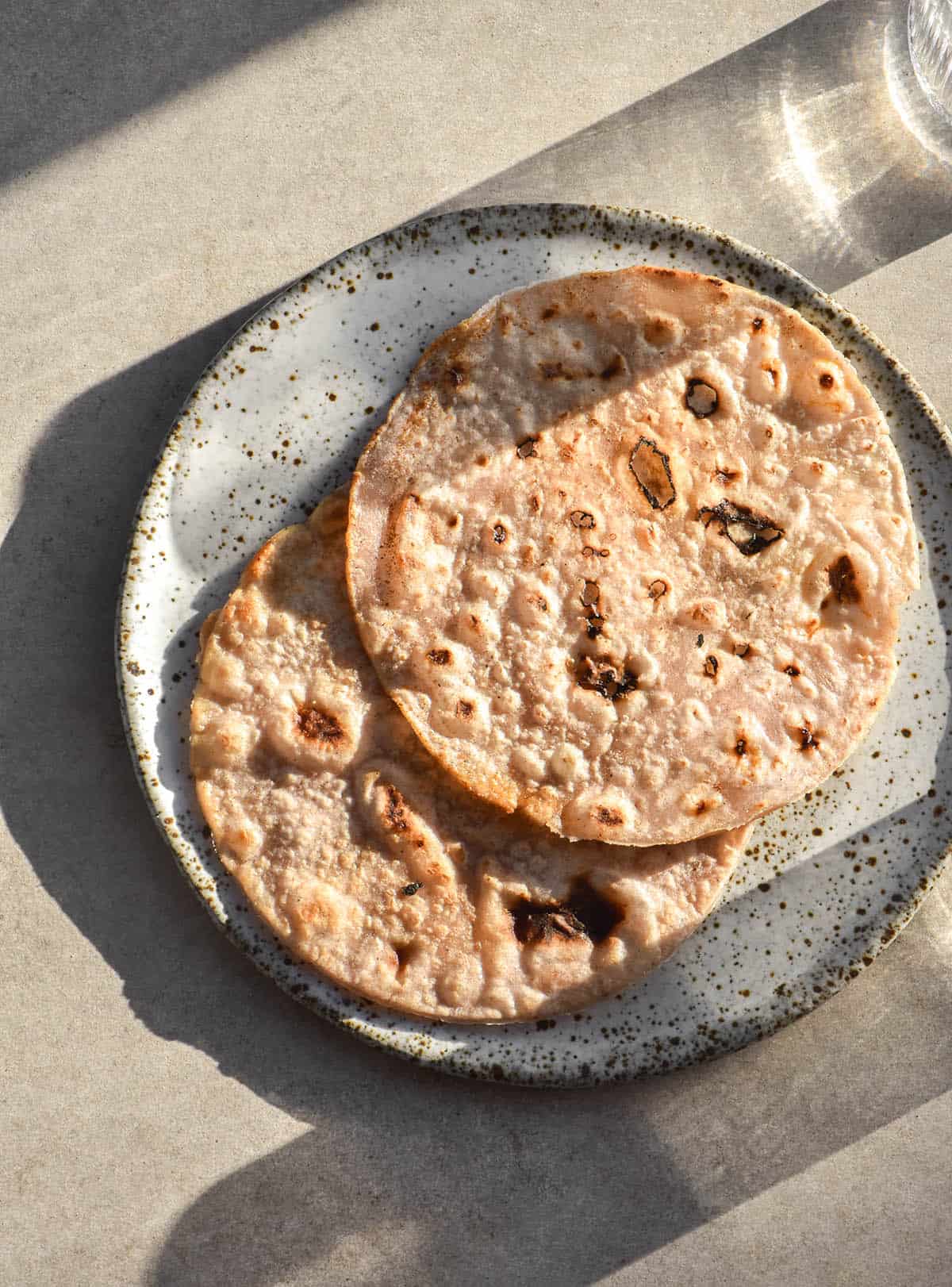 An aerial, sunlit photo of two cassava flour tortillas on a white speckled ceramic plate. The plate sits atop a sunlit stone backdrop and a glass of water sits in the top right and bottom corners, creating a shadow across the image
