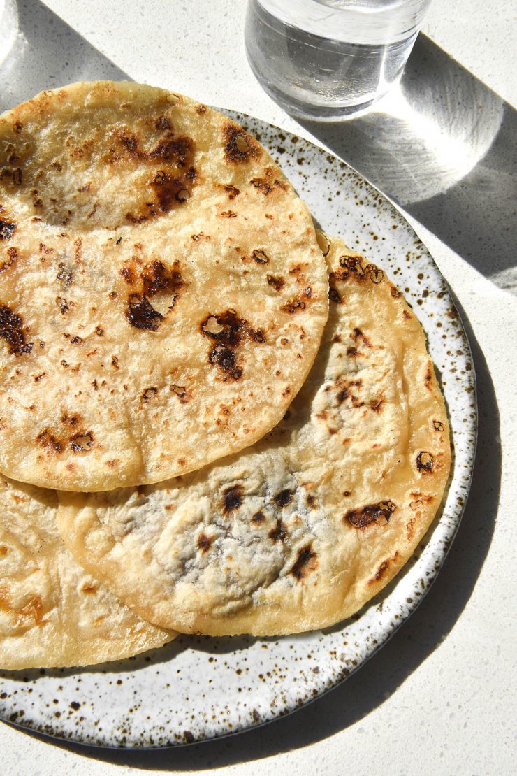 An aerial sunlit view of three gluten free wraps atop a white speckled ceramic plate on a white benchtop. Glasses of water sit in the top left and right hand corners, creating a light and shadow pattern across to the right of the image