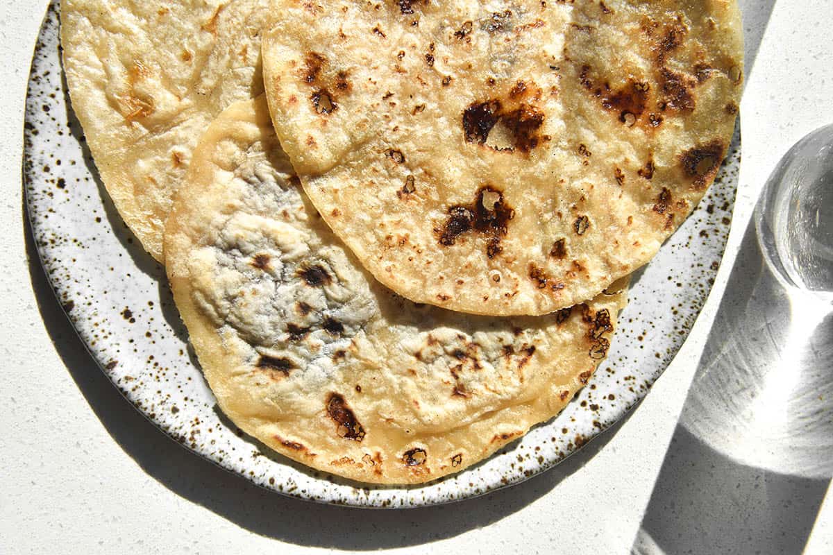 A sunlit image of three grain free cassava flour tortillas atop a white speckled plate on a white stone benchtop. A glass of water sits to the right of the tortillas and casts a shadow from the top to the bottom right of the image.