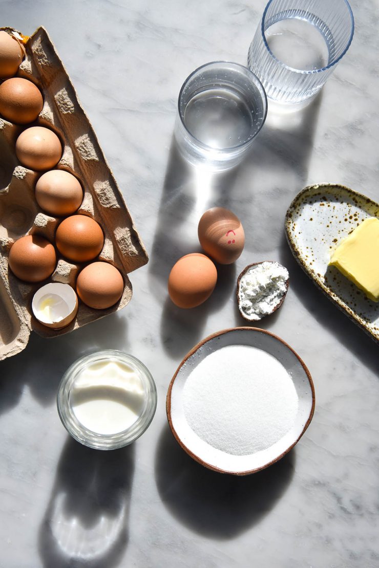 Baking ingredients on a brightly sunlit white marble table. Two glasses of water sit in the top of the image and cast a pattern down across the baking ingredients