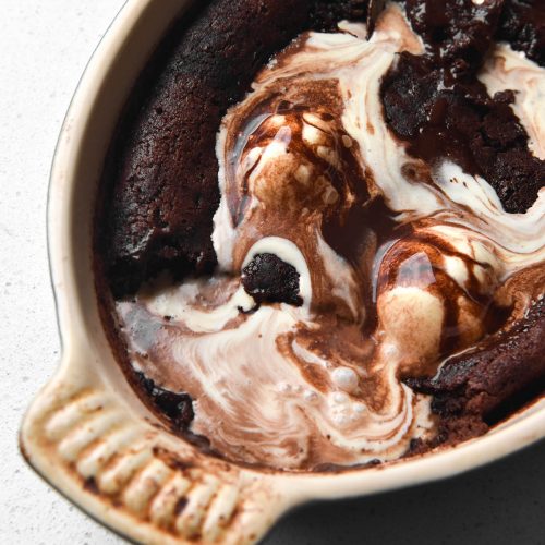 An aerial view of a gluten free chocolate self saucing pudding in an oval shaped baking dish on a white bench top. A few scoops of the pudding have been taken away and melting vanilla ice cream seeps into the pudding.