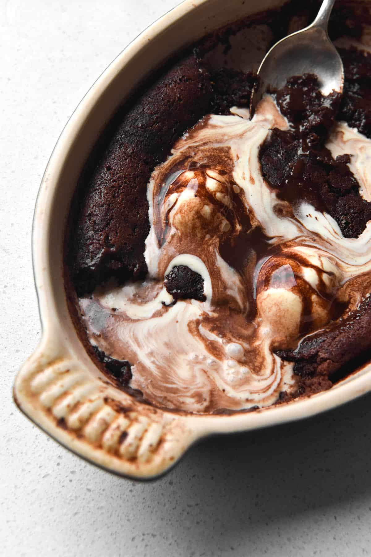 An aerial view of a gluten free chocolate self saucing pudding in an oval shaped baking dish on a white bench top. A few scoops of the pudding have been taken away and melting vanilla ice cream seeps into the pudding.