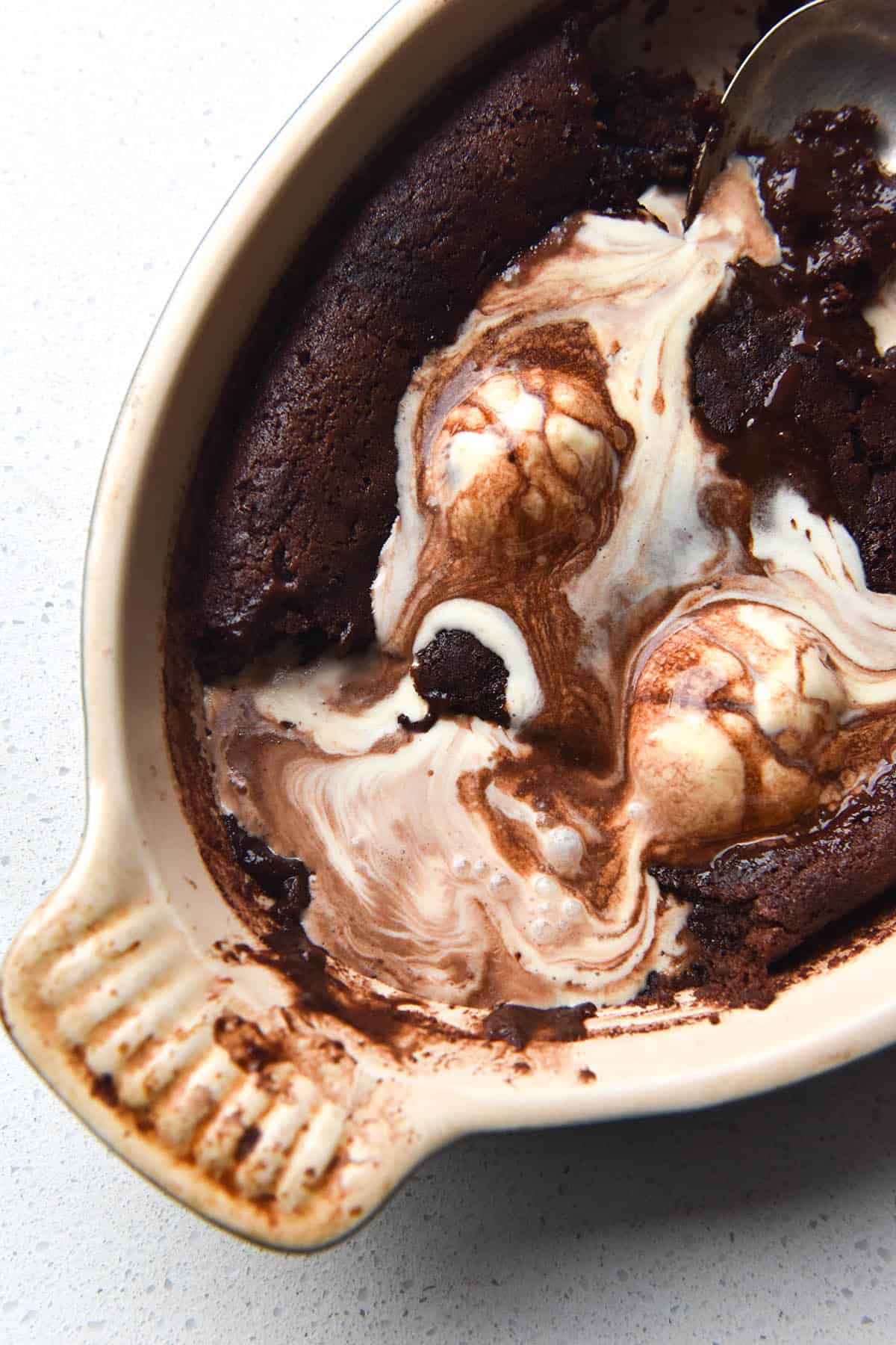 An aerial view of a gluten free chocolate self saucing pudding in an oval shaped baking dish on a white bench top. A few scoops of the pudding have been taken away and melting vanilla ice cream seeps into the pudding.