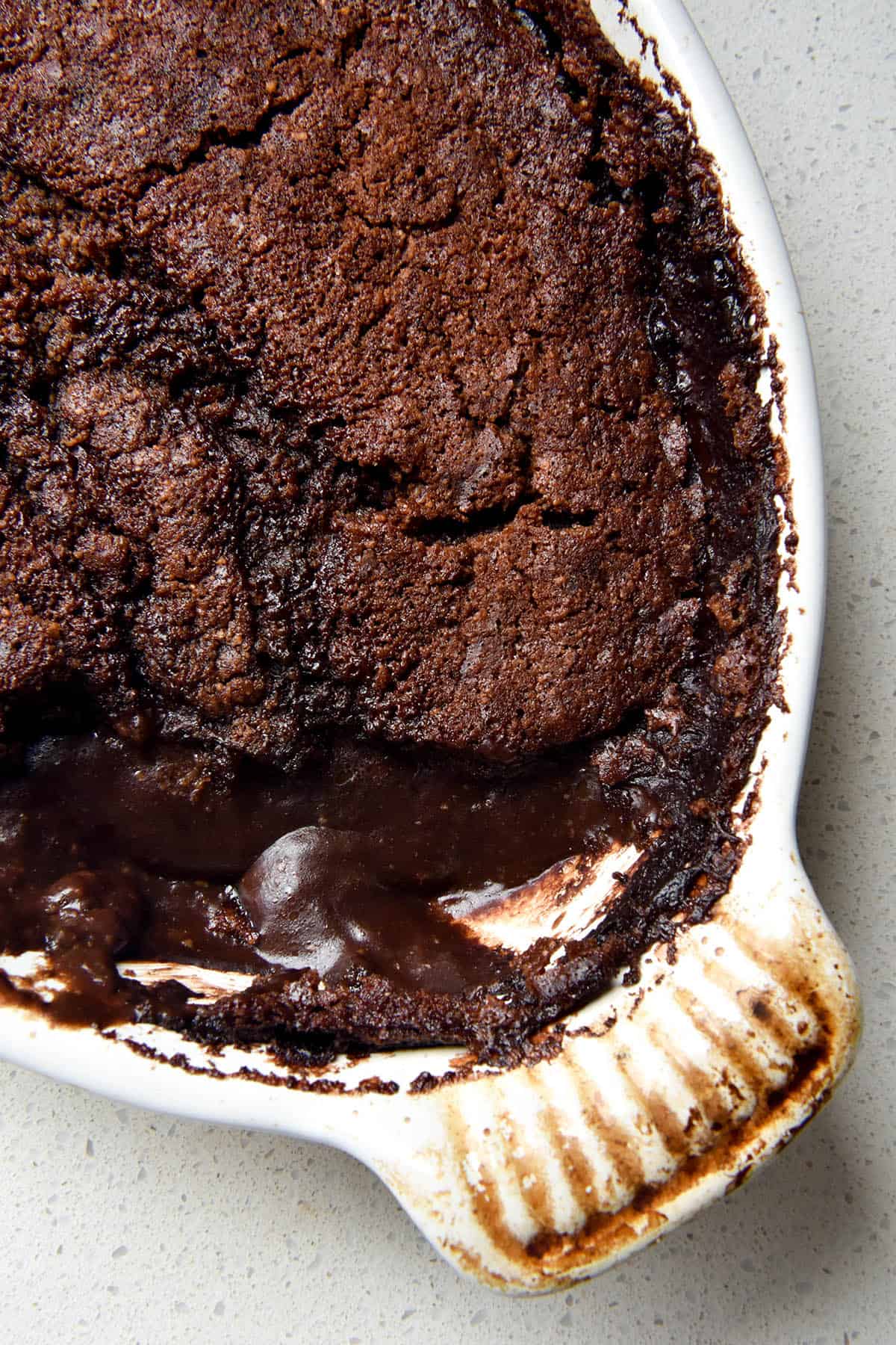 An aerial close up view of a gluten free chocolate self saucing pudding in an oval shaped white baking dish with fluted handles. Spoonfuls of the pudding have been removed and the pudding-like chocolate sauce oozes out from underneath